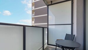 Un balcó amb taula i cadires, una partició de vidre, i un edifici amb balcons al fons.