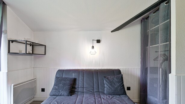 A modern bedroom with a grey sofa, two patterned pillows, a wall-mounted light, and a radiator. - Modern Apartment in Manigod-La Clusaz with Mountain View, Shared Pool & Wi-Fi (Manigod)