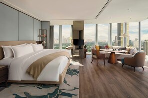 Una habitación de hotel moderna con una cama grande, un sofá, una mesa de comedor y una televisión. La habitación tiene ventanas amplias con vistas a la ciudad.