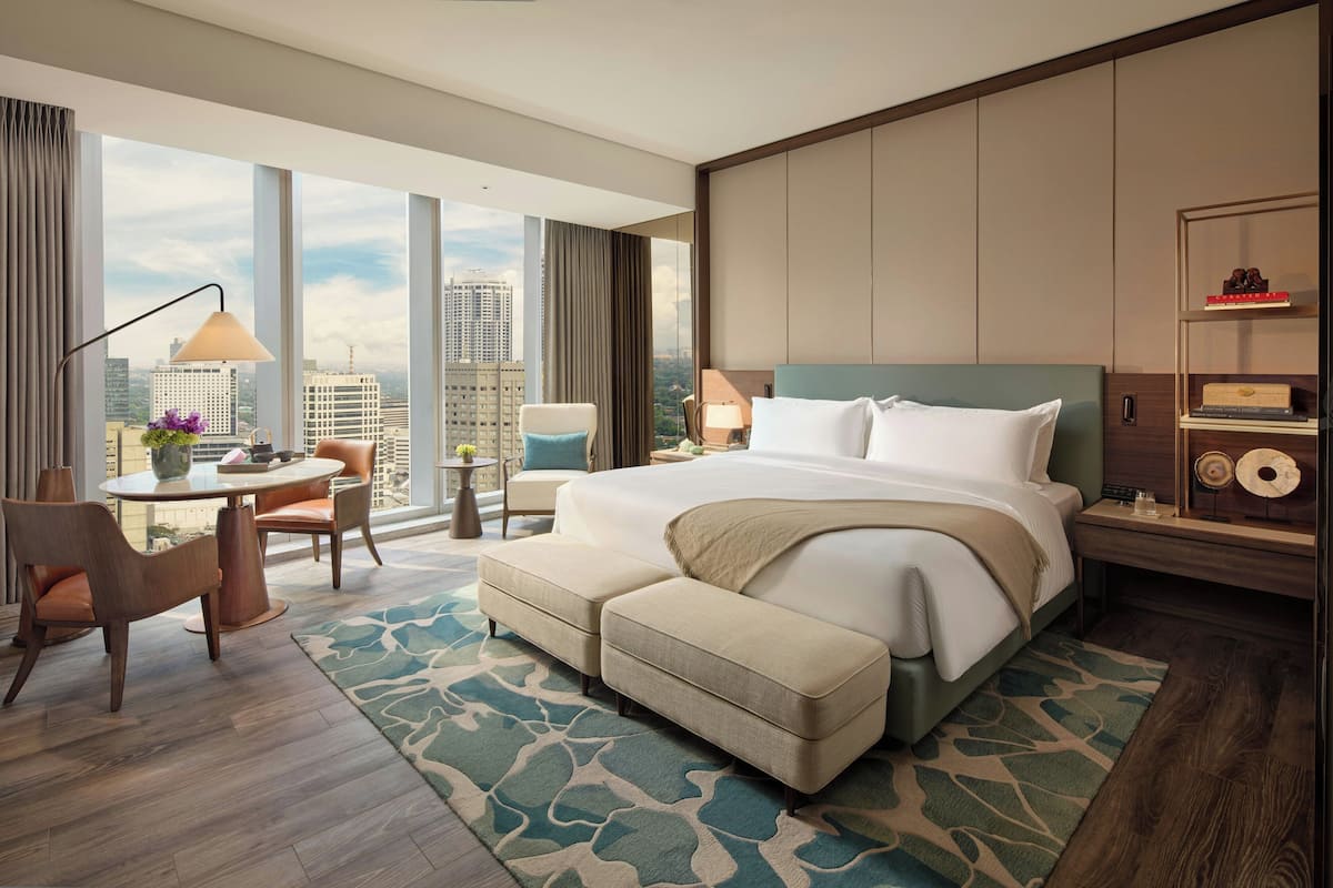 A modern hotel room with a large bed, a desk with a chair, a small table with a lamp, and a view of the city skyline.