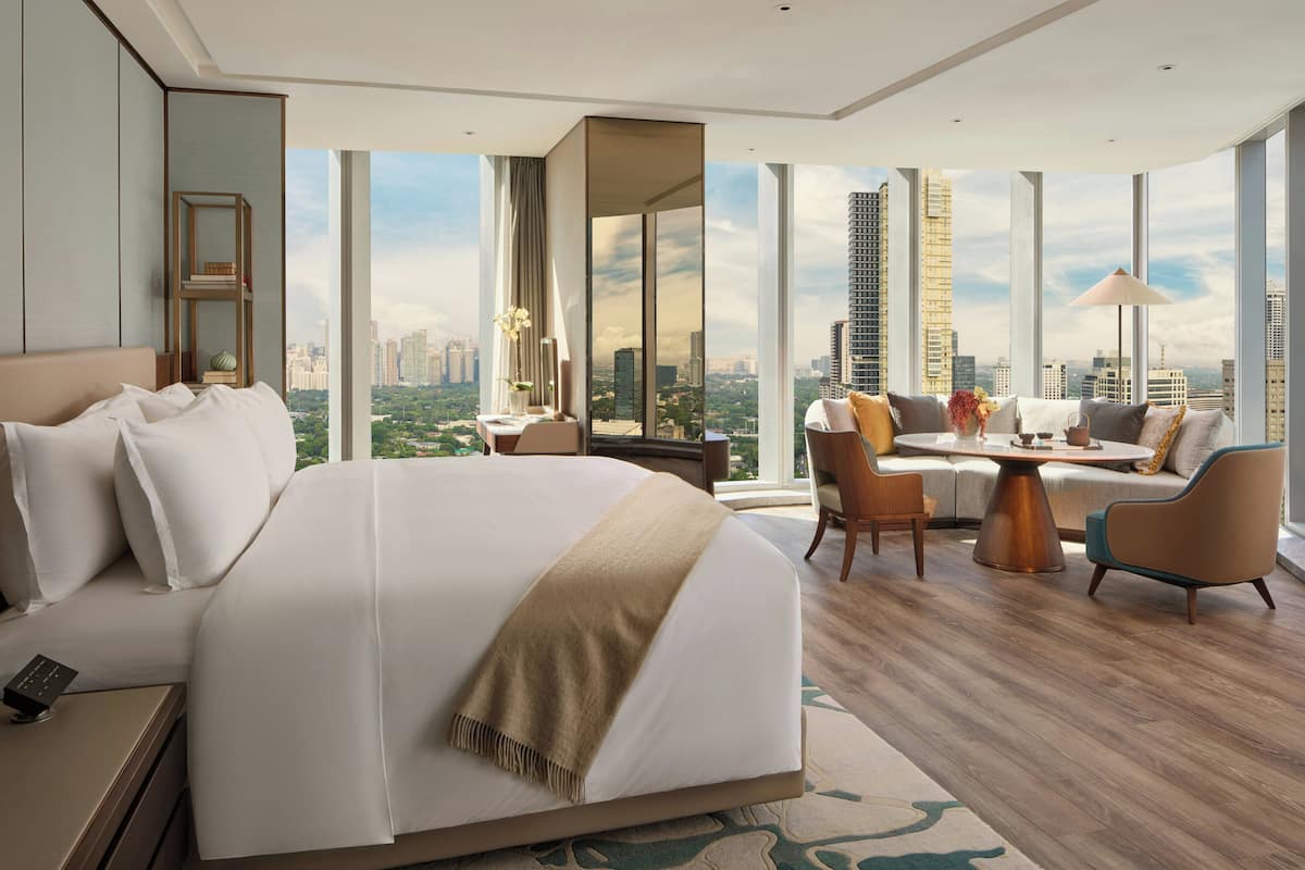 A modern hotel room with a large bed, a sofa, a dining table, and a view of the cityscape.