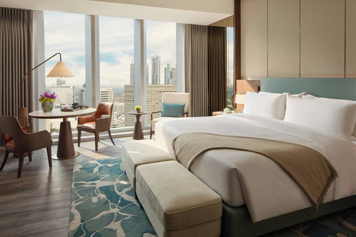 A hotel room with a large bed, a desk with a lamp, two chairs, a small table with a vase, and a view of the cityscape.