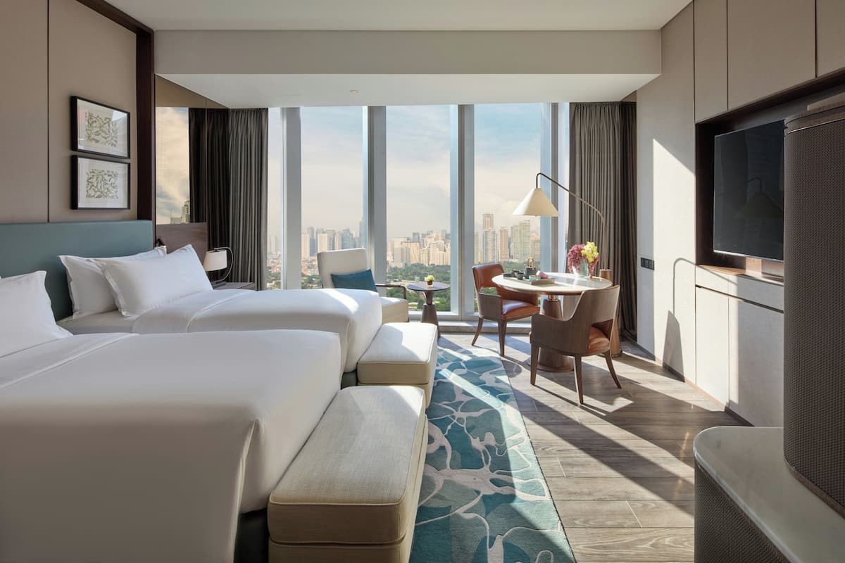 A hotel room with two beds, a desk, a TV, and a city view.