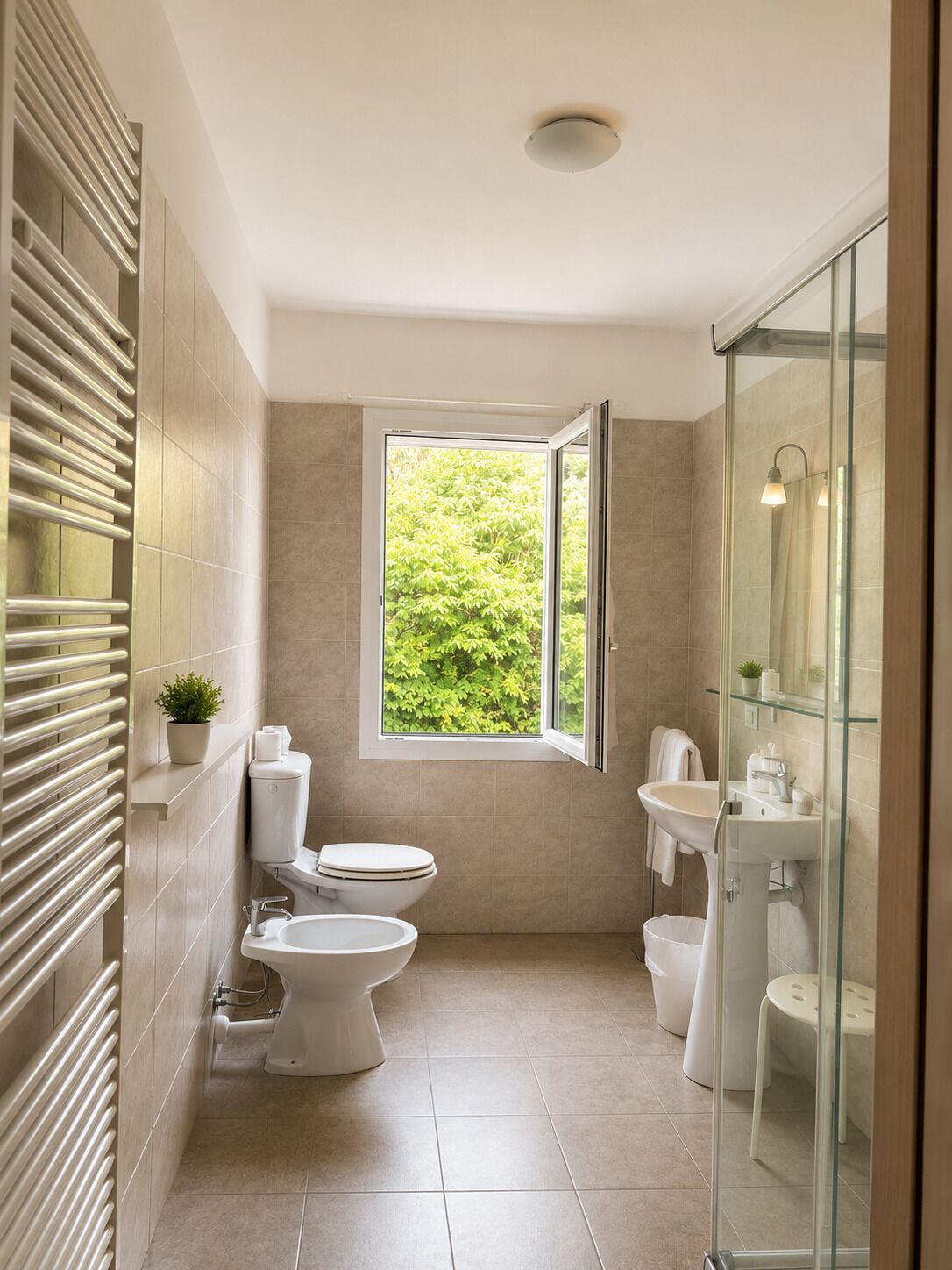 Un baño moderno con inodoro, bidé y lavamanos, cabina de ducha de vidrio y ventana con vistas a la vegetación.