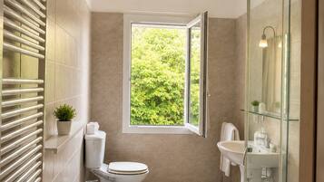 Un baño moderno con inodoro, bidé y lavamanos, cabina de ducha de vidrio y ventana con vistas a la vegetación.