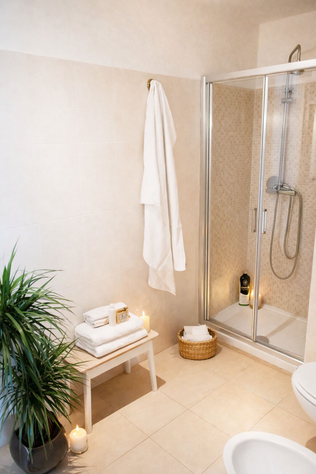 A bathroom with a glass shower enclosure, a white sink, a toilet, a small table with towels, and a potted plant.