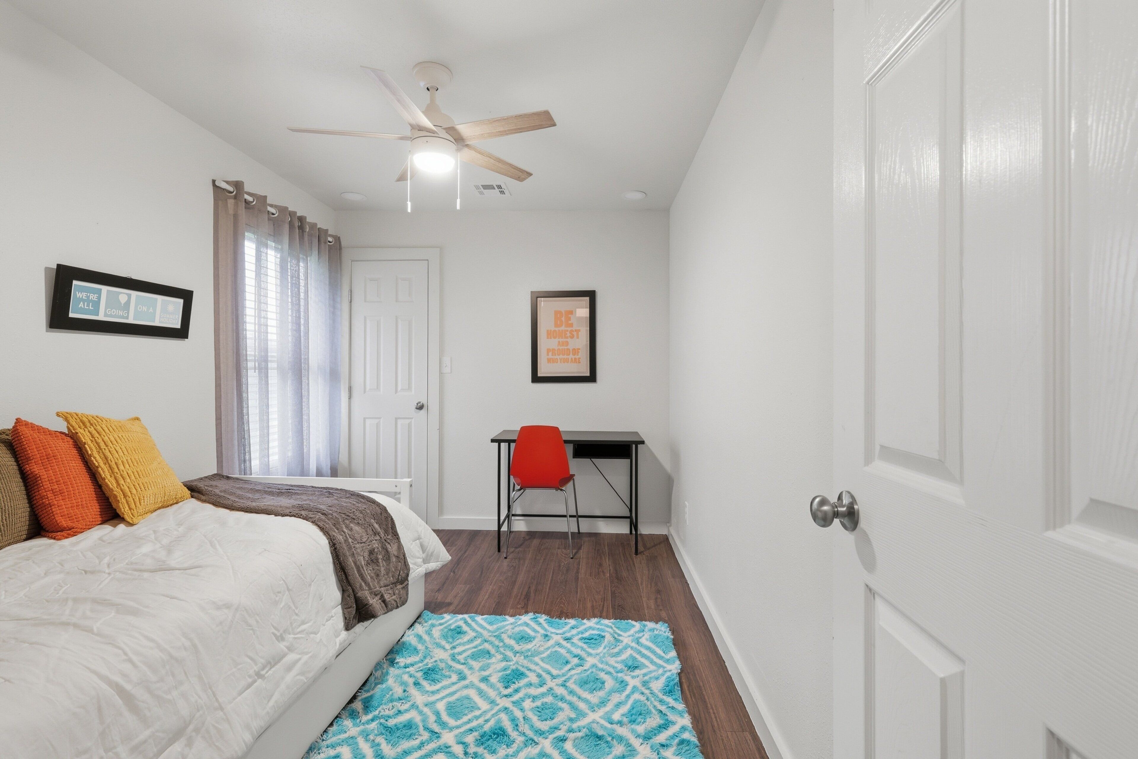 A bedroom with a bed, a desk, a chair, a ceiling fan, and a door.
