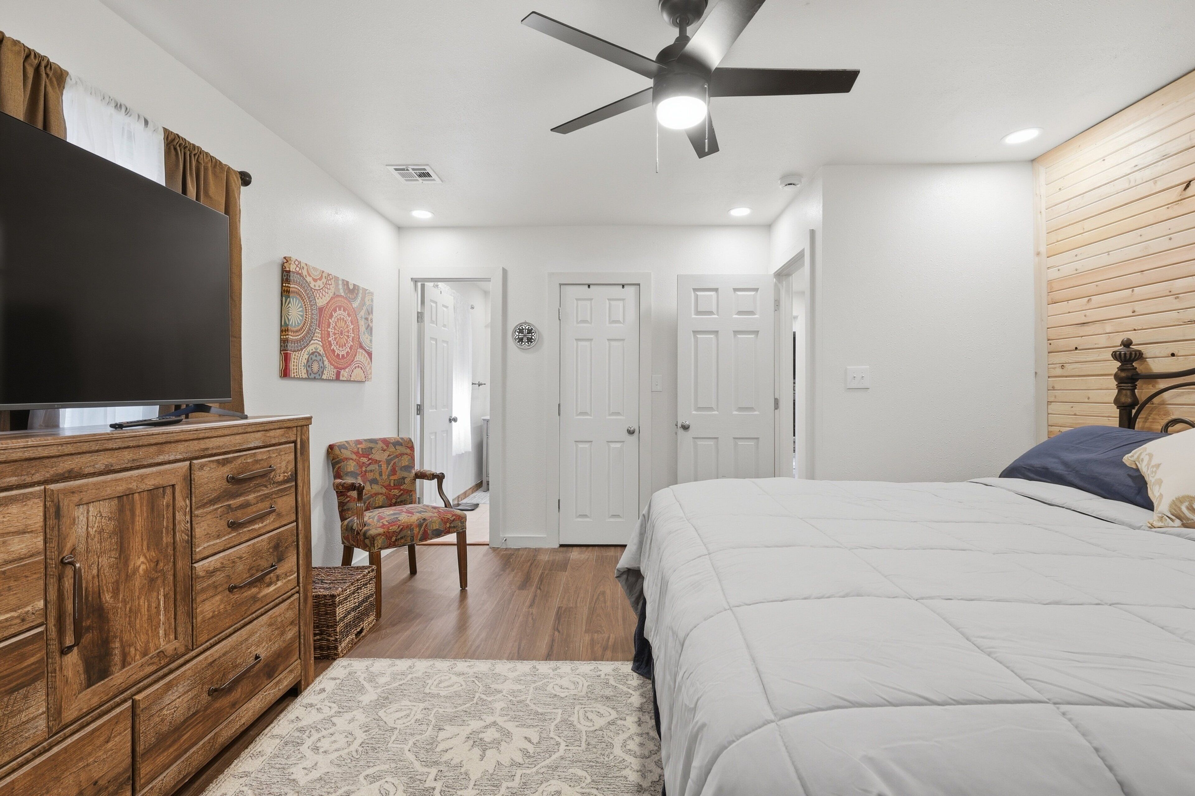 A bedroom with a bed, dresser, television, ceiling fan, and patterned rug.