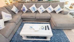 Un sofá modular con almohadones decorativos, una mesa de centro blanca con controles remotos, y una alfombra azul.