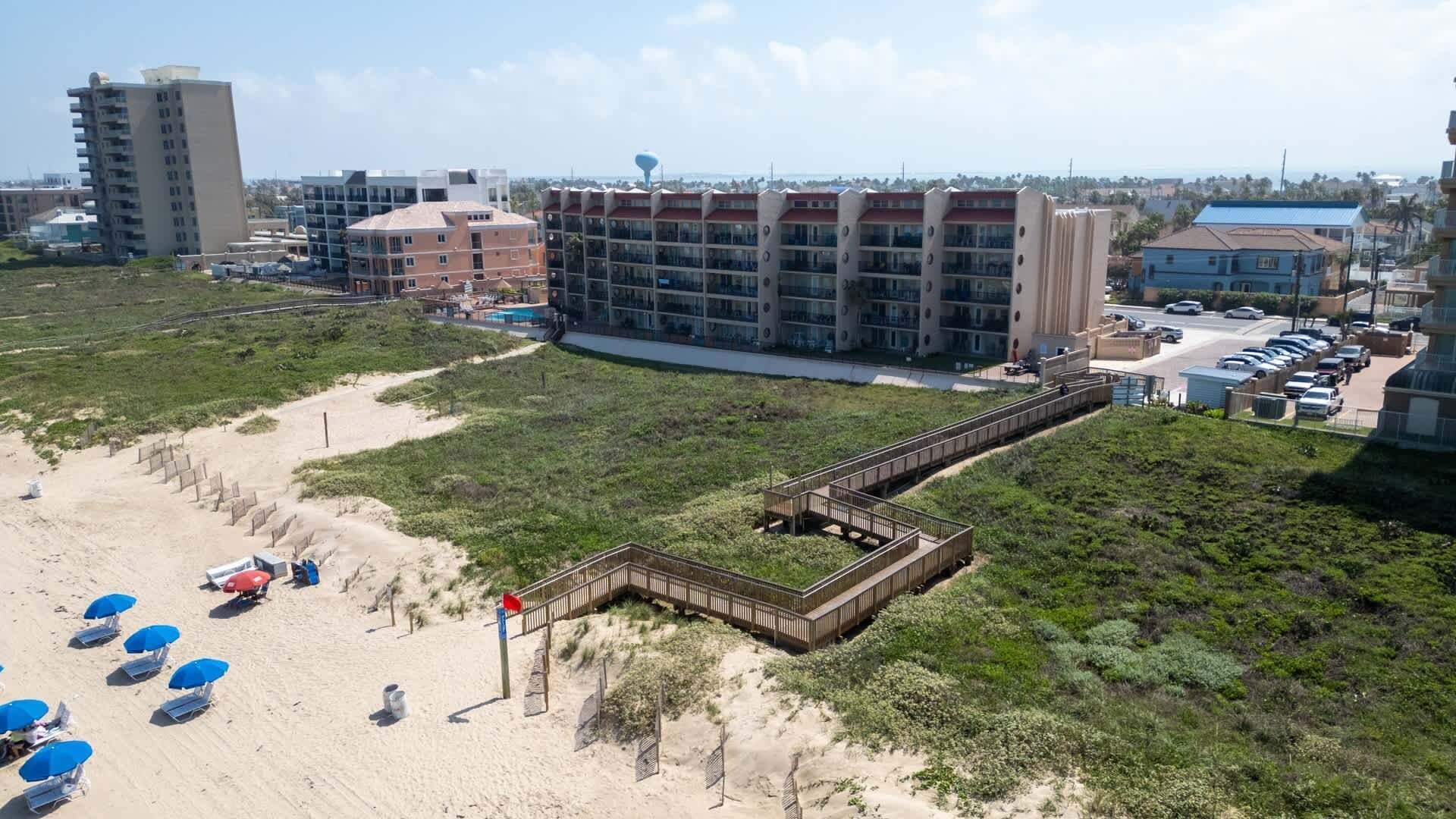 Un hôtel situé sur la côte, avec une promenade en bord de mer menant à la plage, plusieurs bâtiments et un espace de stationnement.