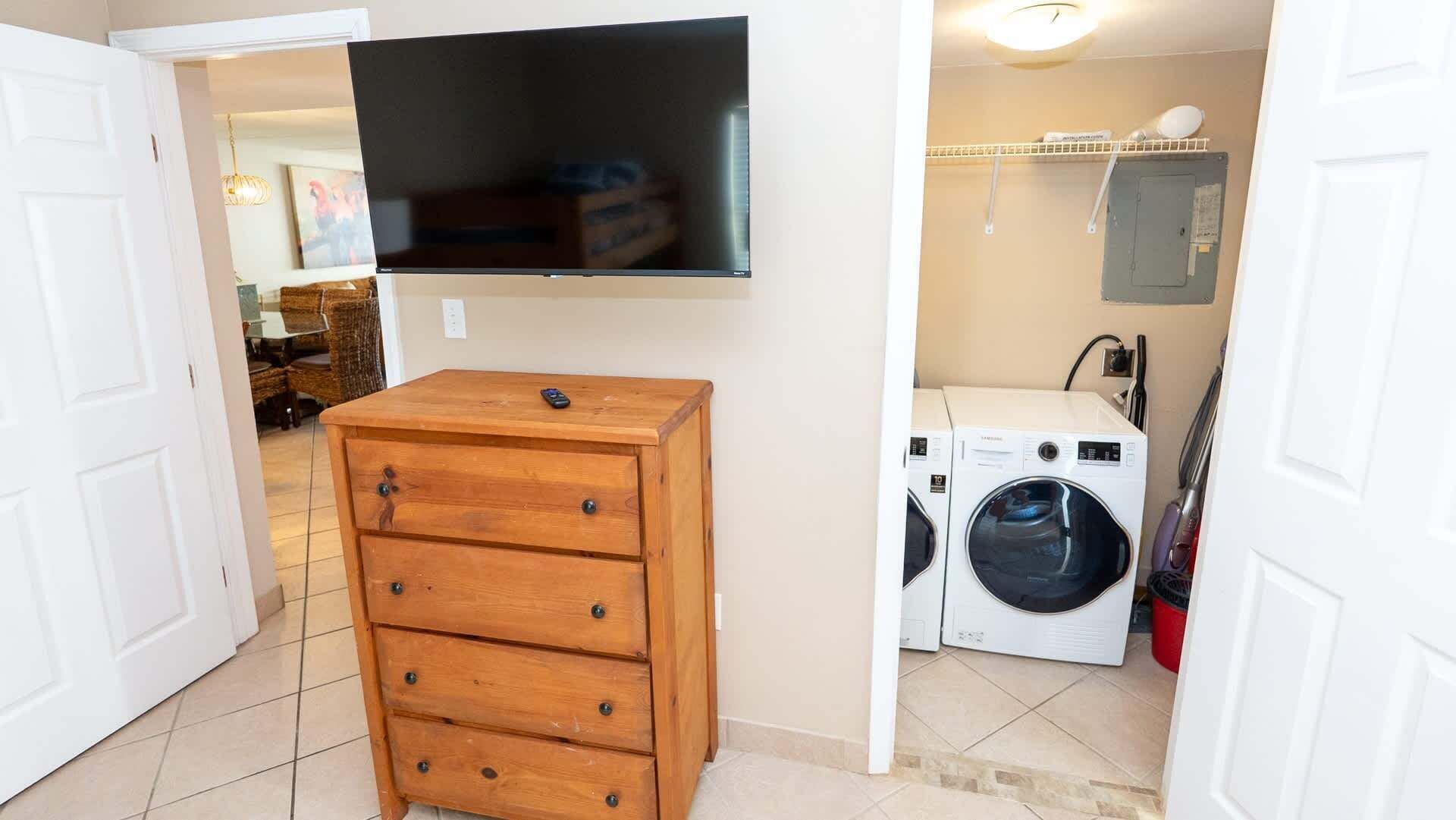 A laundry room with a washing machine and dryer, a wooden dresser, and a television mounted on the wall.