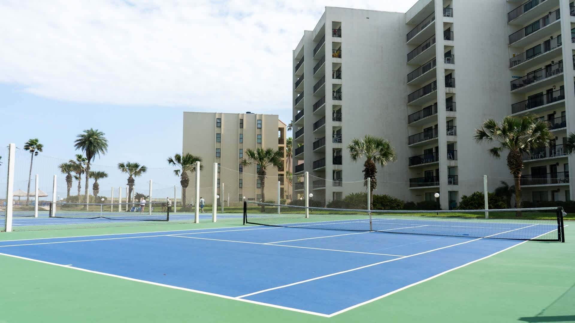 A tennis court with a net, surrounded by palm trees and a multi-story building.