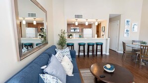 A modern living room with a blue sofa, a dining table, and a kitchen area with bar stools. - Family Condo Pool, Jacuzzi & Bbq Walk To Beach 2 Bedroom Apts (South Padre Island)