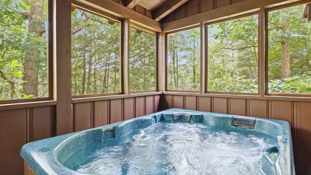 Un bain à remous dans une pièce aux murs en bois et aux grandes fenêtres donnant sur une forêt.