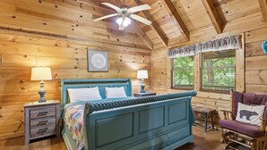 Une chambre dans une cabane en bois, comprenant un lit, deux tables de chevet, une chaise, un ventilateur de plafond et une fenêtre avec des volets.