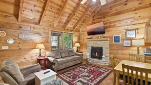 L’intérieur chaleureux d’un chalet avec une cheminée en pierre, des murs en bois, un canapé, une table à manger et un téléviseur fixé au mur.