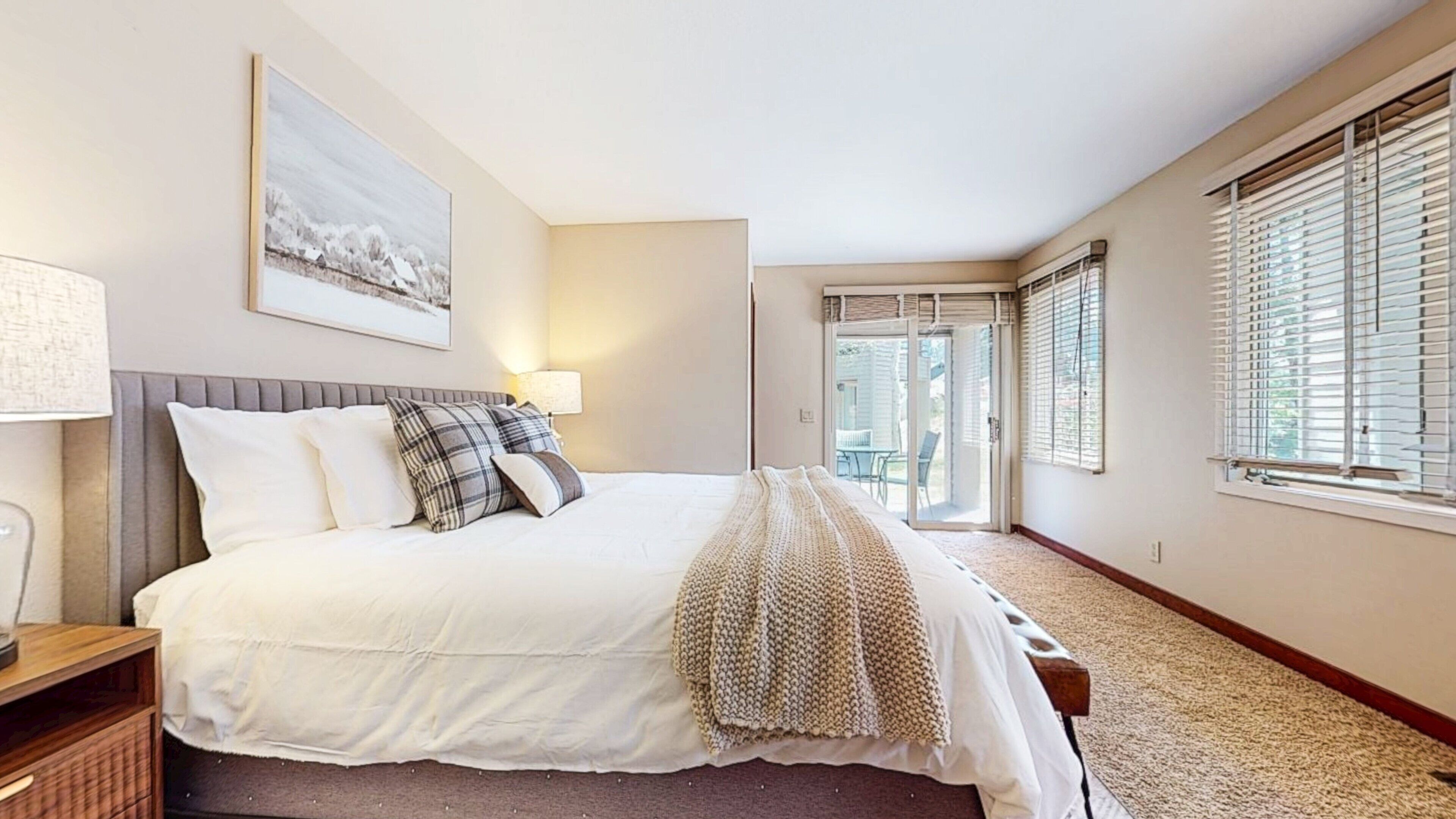 A bedroom with a bed, a bedside table, a lamp, a window with blinds, and a door leading to a balcony.