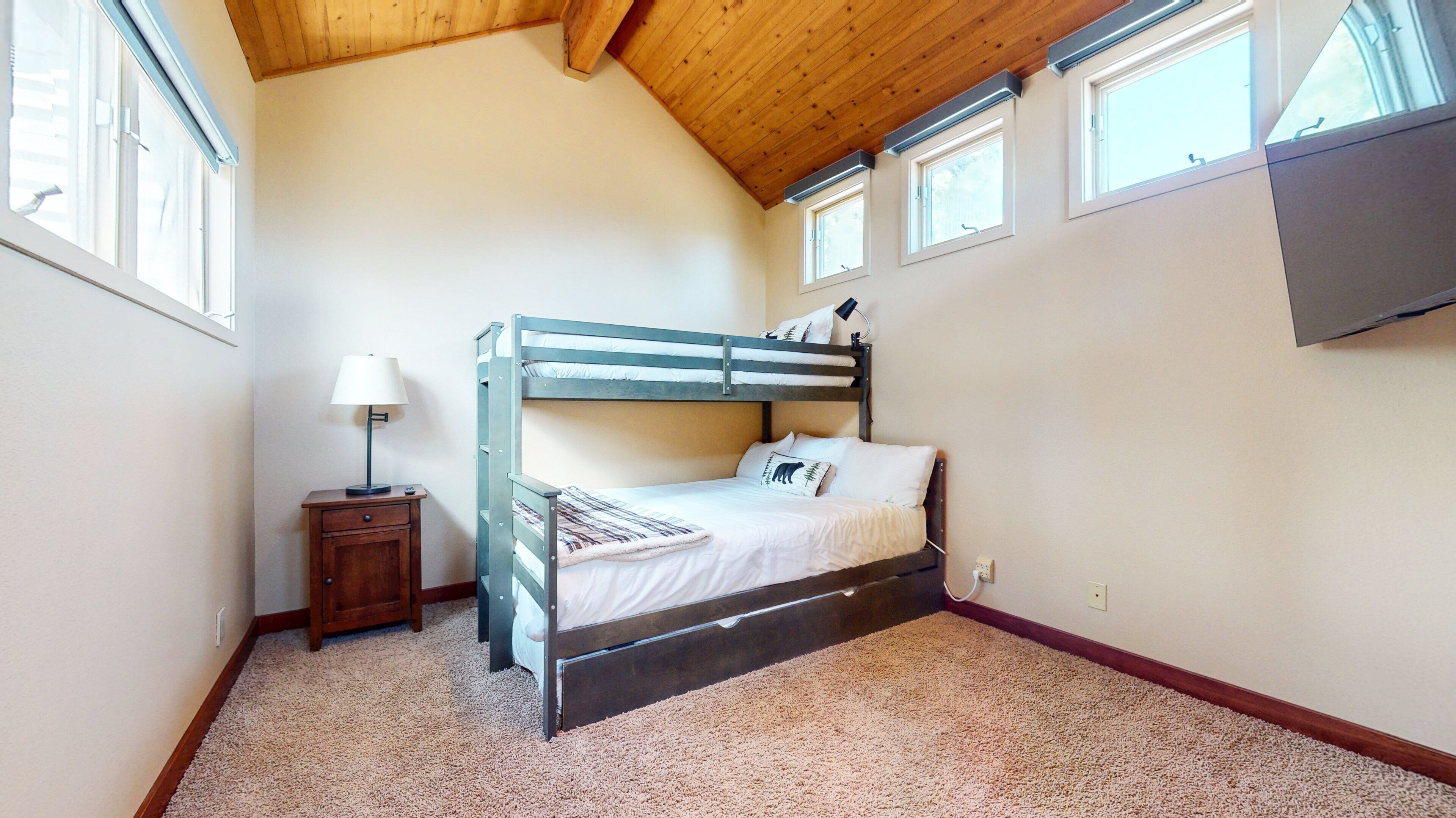 A bunk bed room with a wooden ceiling, a nightstand with a lamp, and a carpeted floor.
