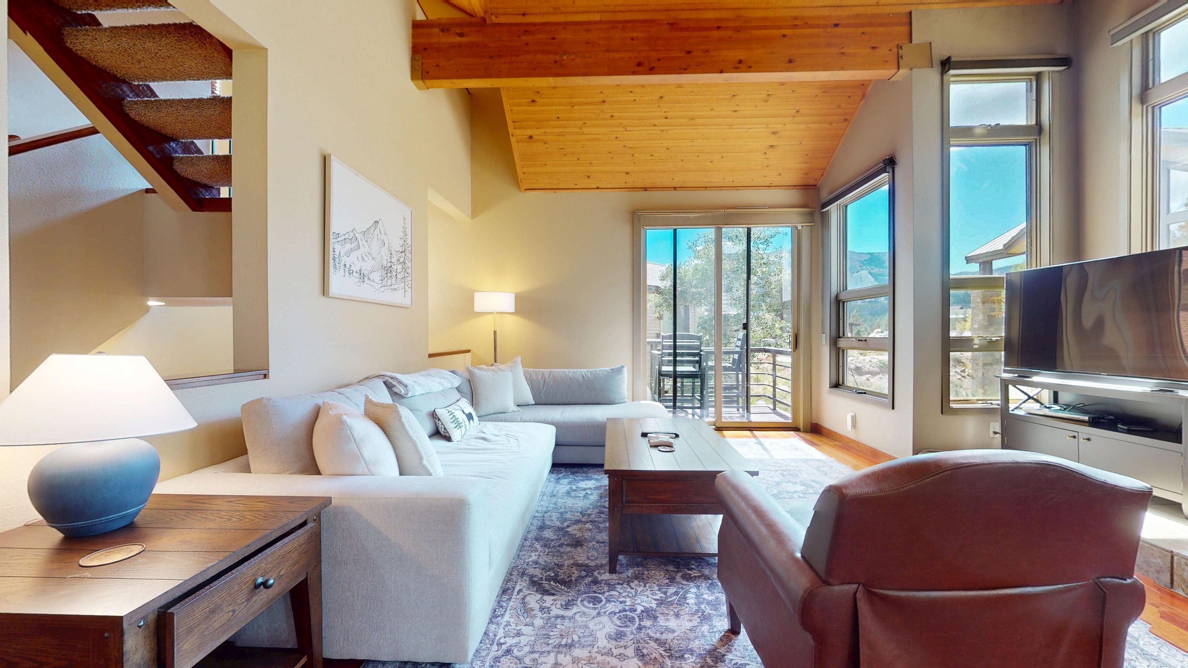 A modern living room with a sofa, coffee table, and TV. There is a staircase on the left and a balcony with a view.