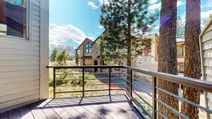 Eine Holzdielen-Terrasse mit Metallgeländer, die einen Wohnbereich mit Steinhäusern und einem Berg im Hintergrund überblickt.