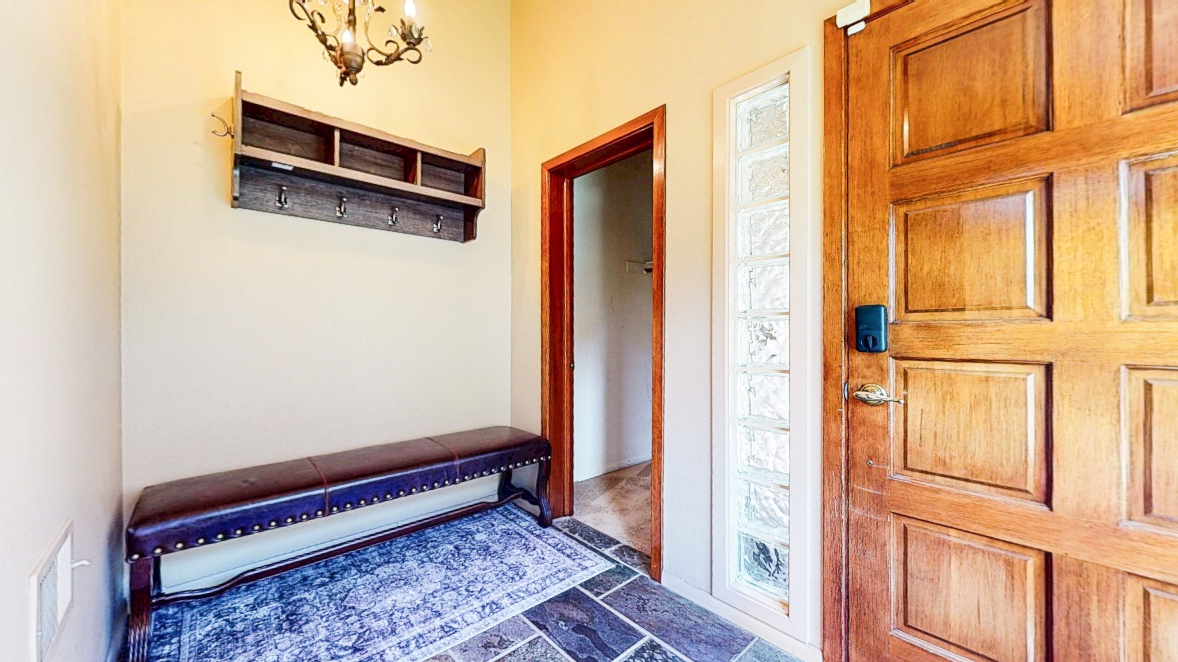 A hallway with a wooden door, a bench, a wall-mounted shelf, and a patterned rug.