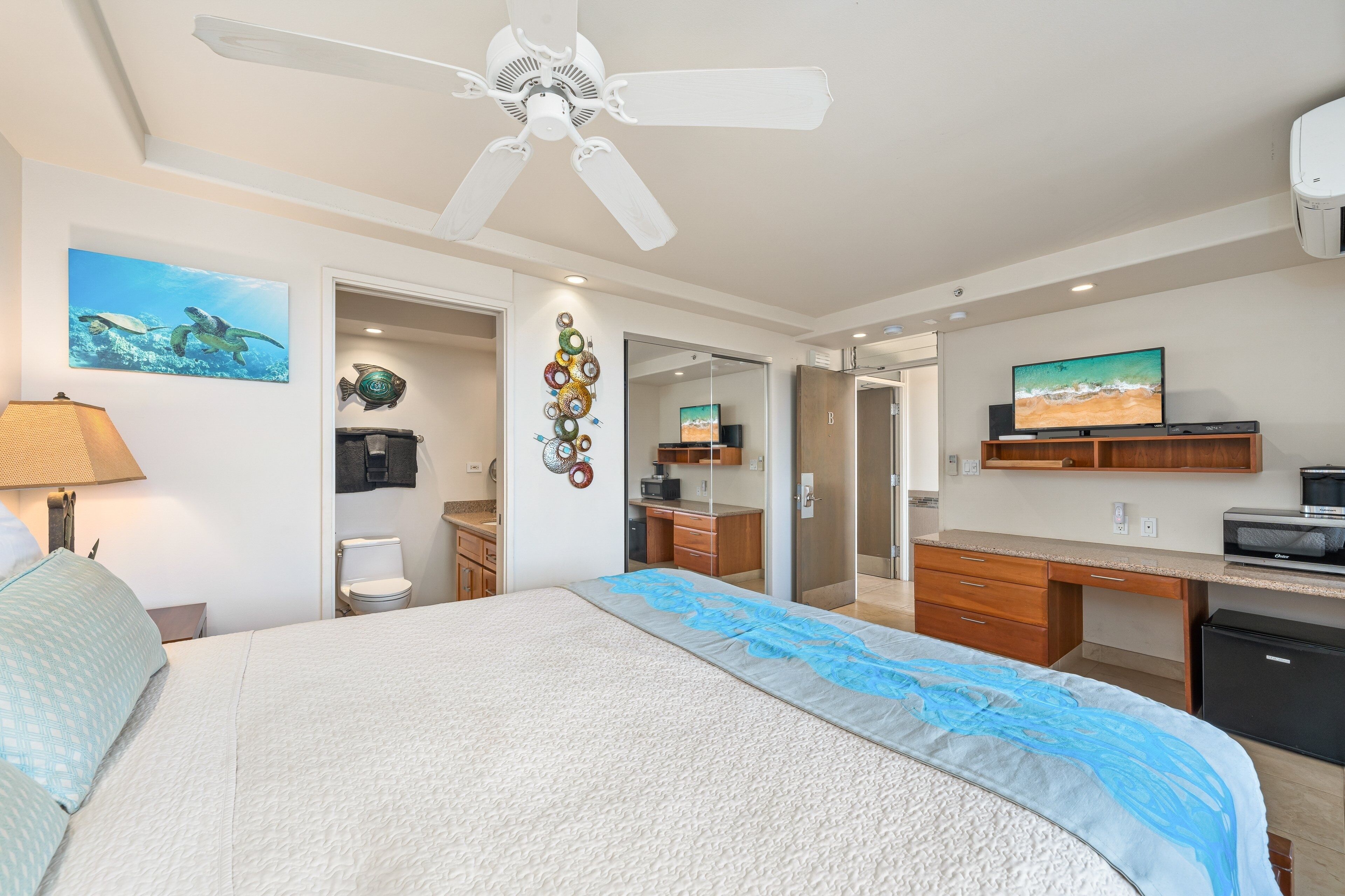 Une chambre à coucher avec un lit, un ventilateur de plafond, une télévision et une salle de bain visible à travers une porte ouverte.