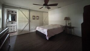 Een slaapkamer met een bed, een plafondventilator, een nachtkastje, een kapstok en een deur naar een andere kamer.