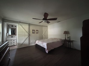 A bedroom with a bed, a ceiling fan, a nightstand, a dresser, and a door leading to another room. - Little Joe’s Cottage (Cocoa)