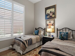 A bedroom with two beds, a window with blinds, a nightstand with a lamp, and artwork on the wall. - Casa Aviano 4 Bedroom Home (Phoenix)