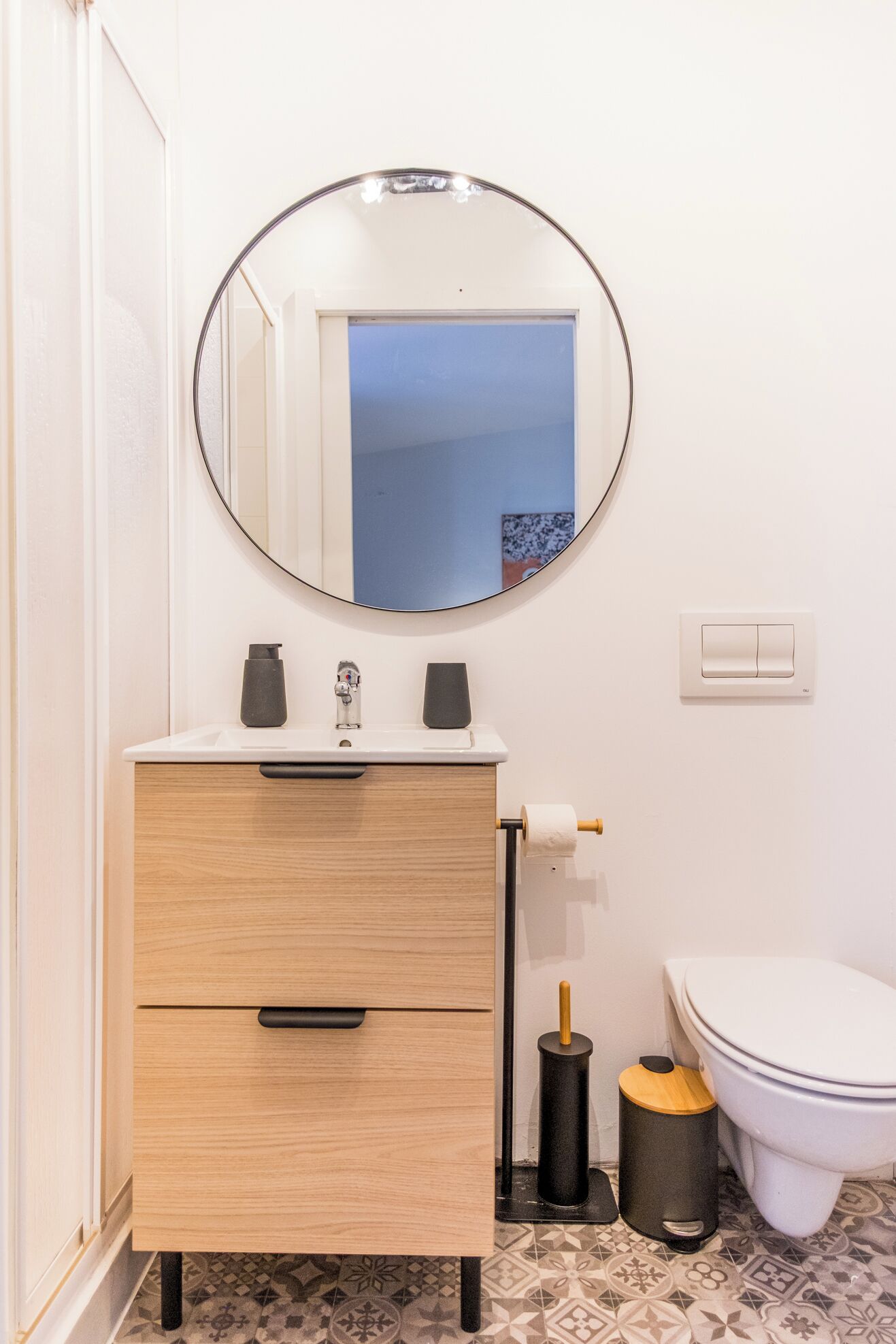 Un baño con tocador de madera, espejo redondo, inodoro y cubo de basura.