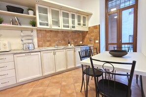 A kitchen with white cabinets, a dining table with chairs, a window with blinds, and a tiled floor. - Old Ghetto Charm. Sleeps 5. Ancient Ruins View (Rome)
