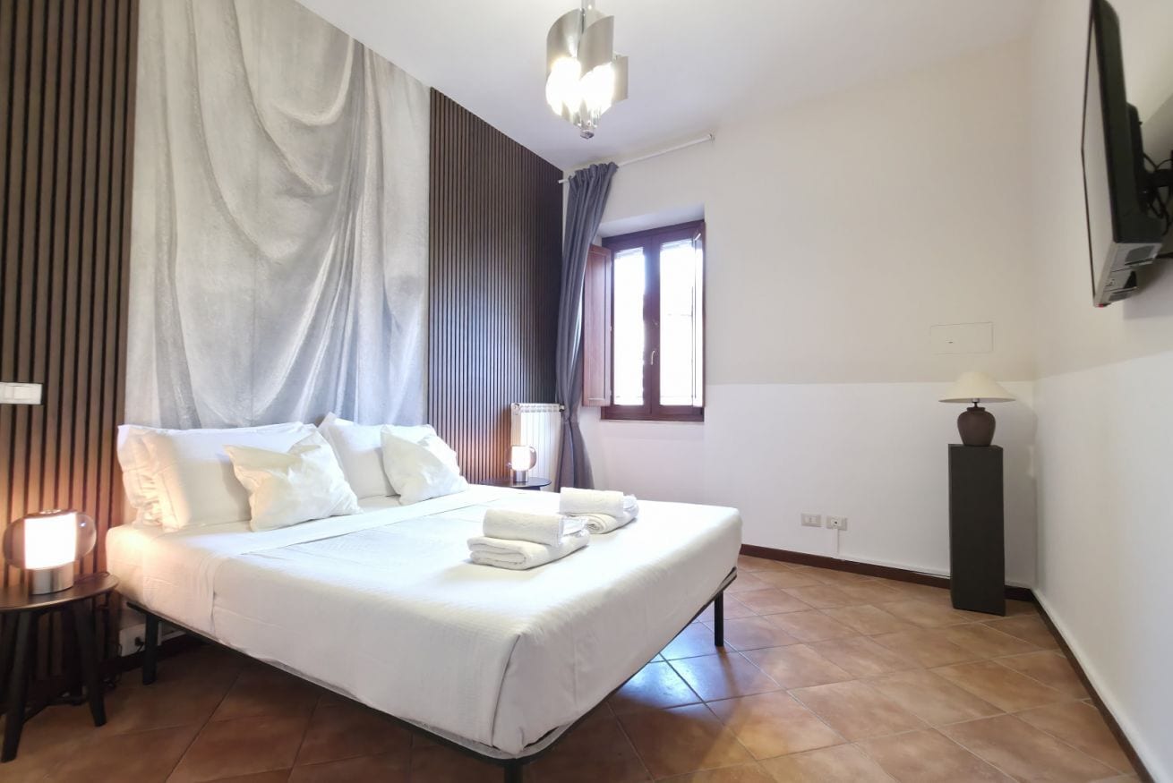 A hotel room with a bed, a nightstand, a lamp, a window, and a wall-mounted TV.