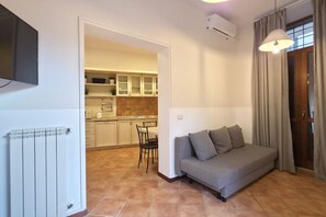 A living room with a sofa, a TV, a radiator, a kitchen in the background, and a window with curtains. - Old Ghetto Charm. Sleeps 5. Ancient Ruins View (Rome)