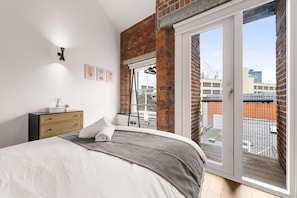 Ein Schlafzimmer mit Bett, Kommode, Fenster mit Blick auf Gebäude und einem Balkon.