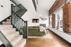 Eine Treppe mit Teppichstufen und einem grünen Geländer, ein grünes Sofa mit Decke, ein an der Wand befestigtes Fernsehgerät und eine Ziegelwand mit Fenstern.