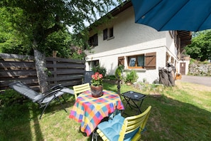 Une terrasse avec une table et des chaises, un pot de fleurs et une maison avec un toit en pente et des volets en bois.