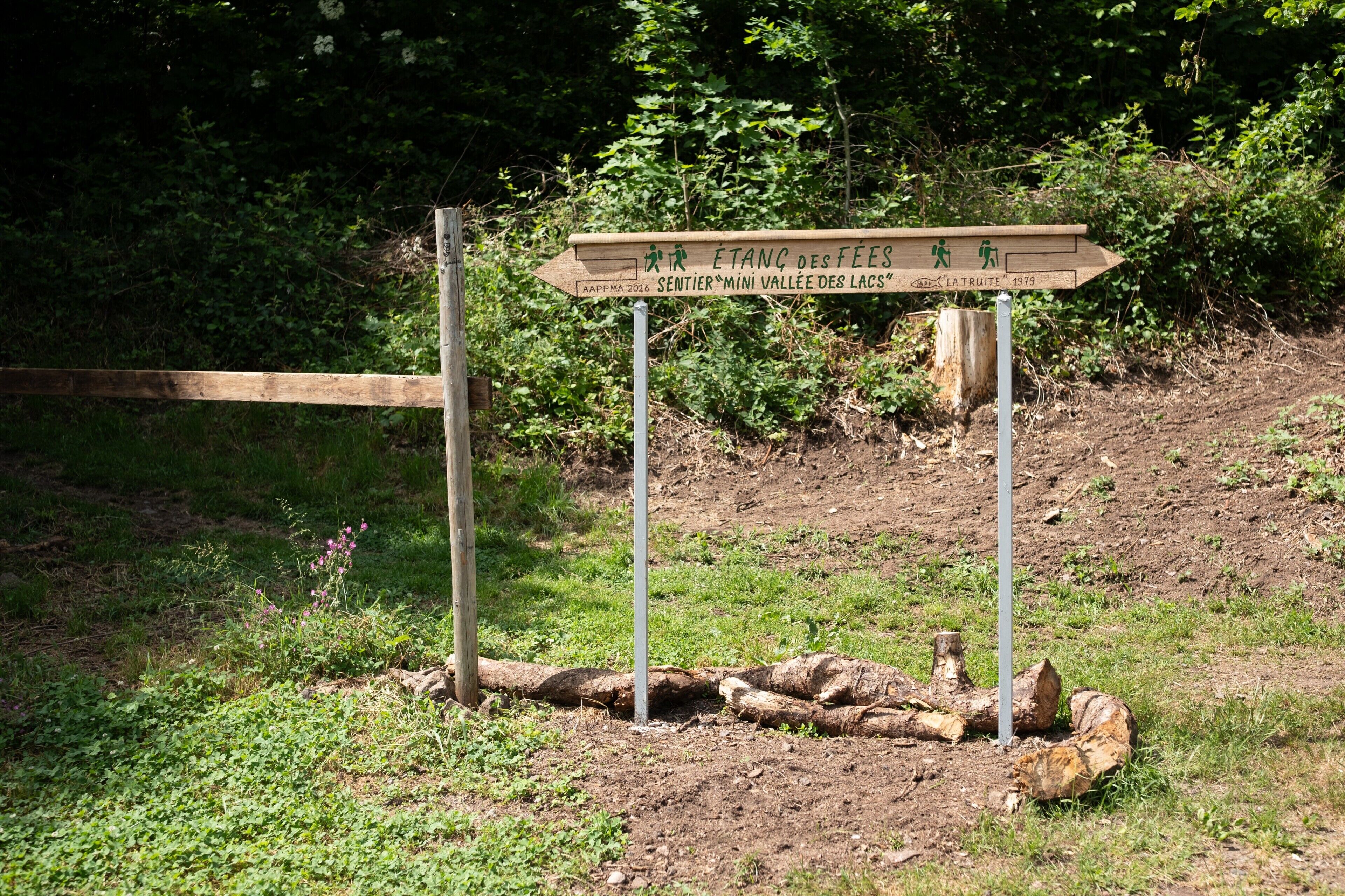 Un panneau indicateur en bois, avec des inscriptions en français, entouré de verdure et bordé d’un chemin de terre.