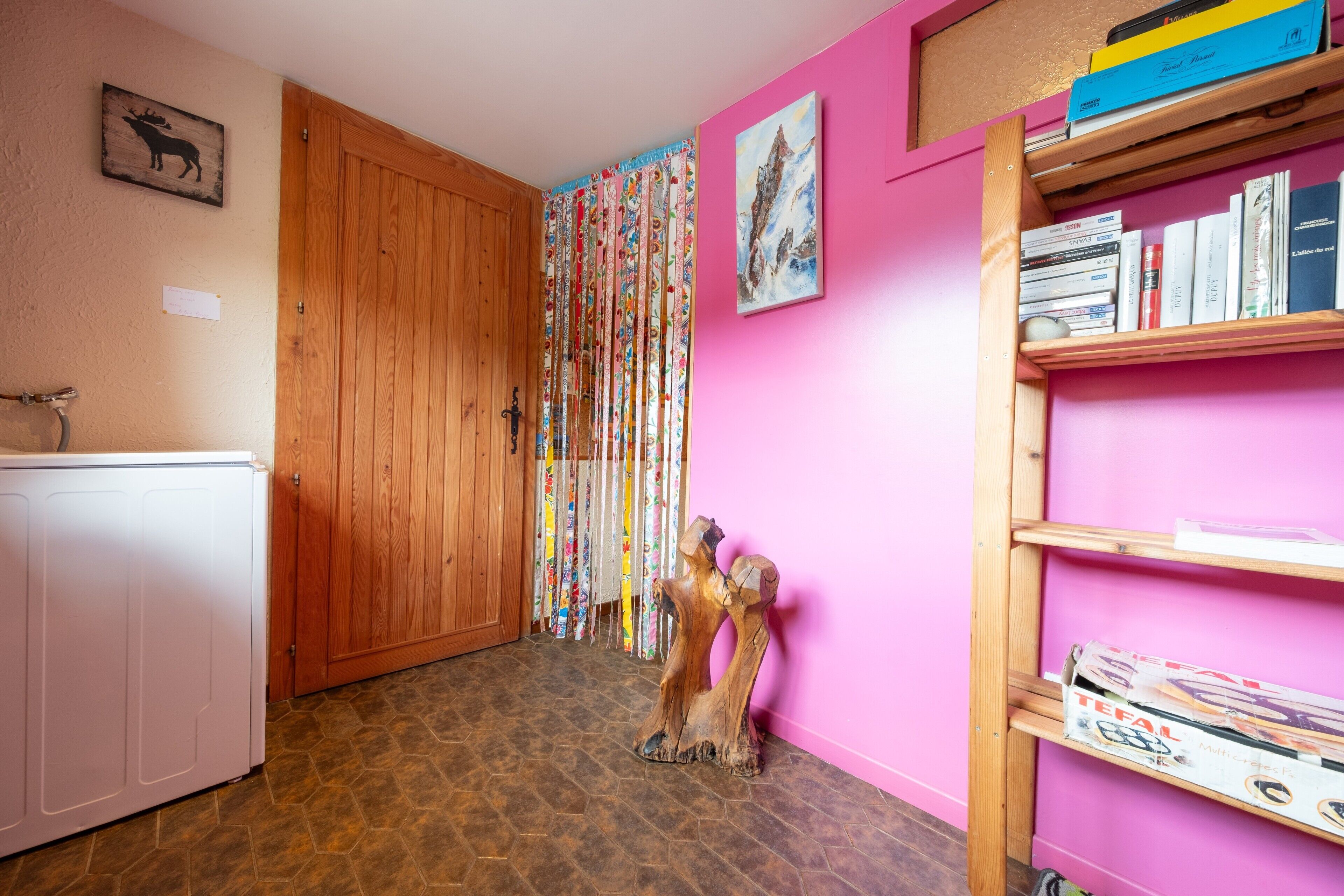 Ein Raum mit einer Holztür, einem weißen Gerät, einer rosa Wand, einem Bücherregal und einer dekorativen Holzskulptur.