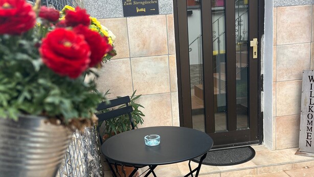 Un patio con mesa y sillas, una planta en maceta, una puerta y un cartel de bienvenida.