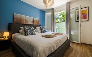 Ein Schlafzimmer mit Bett, Kissen, Nachttisch, Lampe, einem Gemälde und Blick ins Freie.
