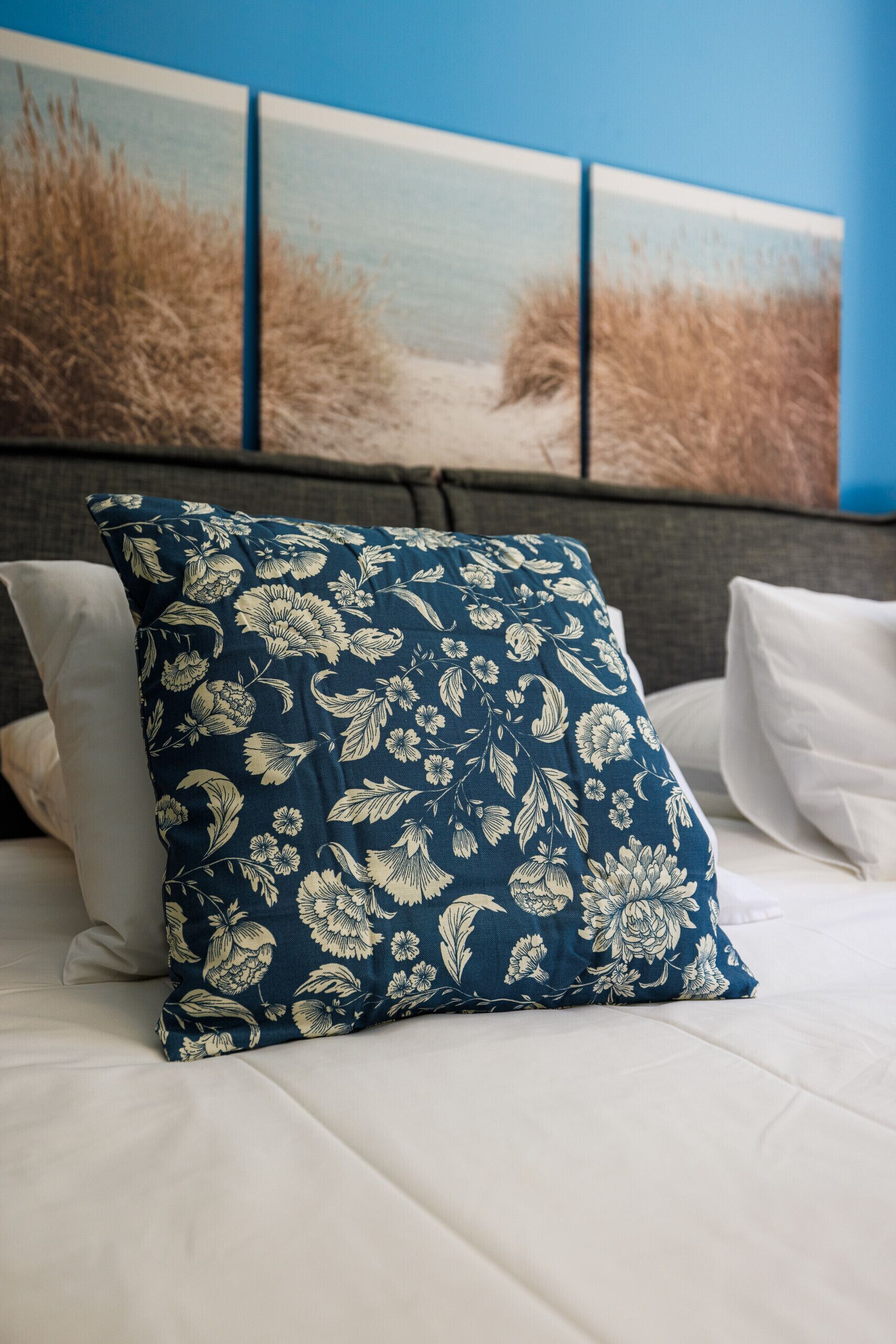A bed with white bedding, a blue floral pillow, and a headboard with a painting of a beach scene.