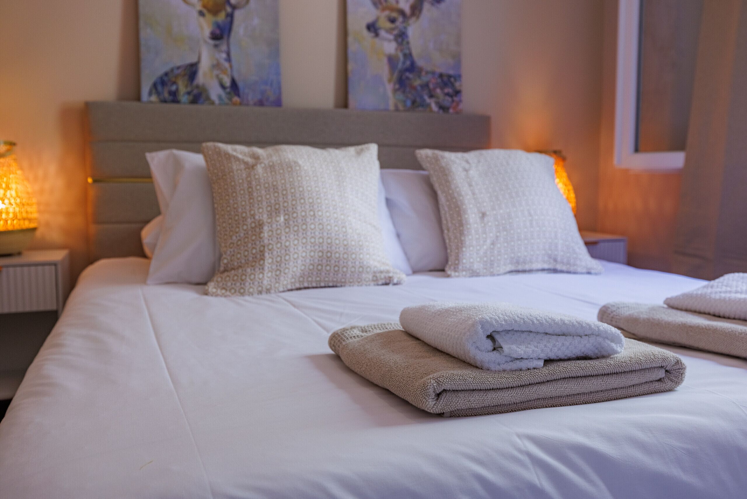 Een net opgemaakt bed met kussens, gevouwen handdoeken, een hoofdbord en wanddecoratie.