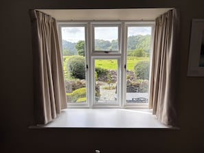 A room with a large window featuring a view of a green landscape with trees and a stone wall. - The Barn at Grasmere (Ambleside)