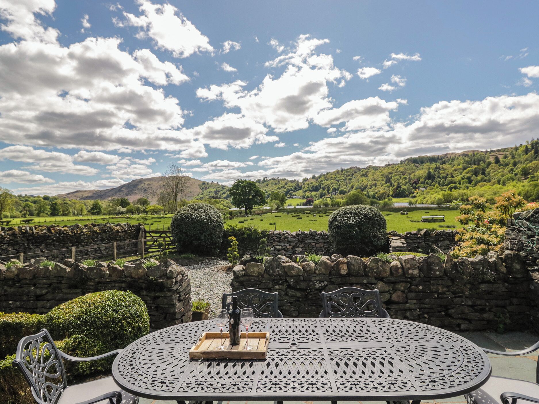 A round metal table with chairs, a bottle, and glasses, set against a scenic backdrop of a green valley and hills under a partly cloudy sky.