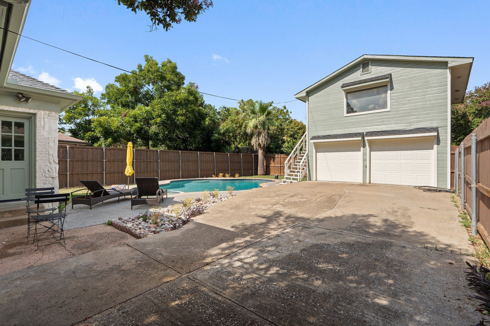 A backyard with a swimming pool, lounge chairs, a bench, a covered patio area, and a garage.