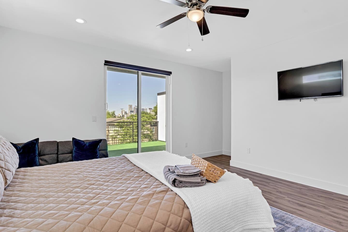 A bedroom with a bed, pillows, a flat-screen TV, a ceiling fan, and a view of the city through a sliding door.