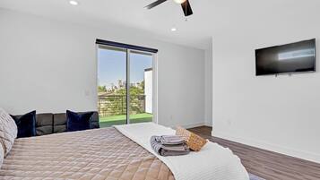 A bedroom with a bed, pillows, a flat-screen TV, a ceiling fan, and a view of the city through a sliding door.