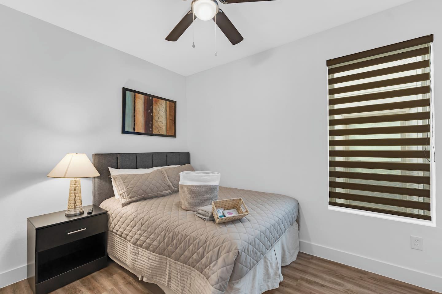 A bedroom with a bed, a nightstand, a lamp, a ceiling fan, a window with blinds, and a painting on the wall.