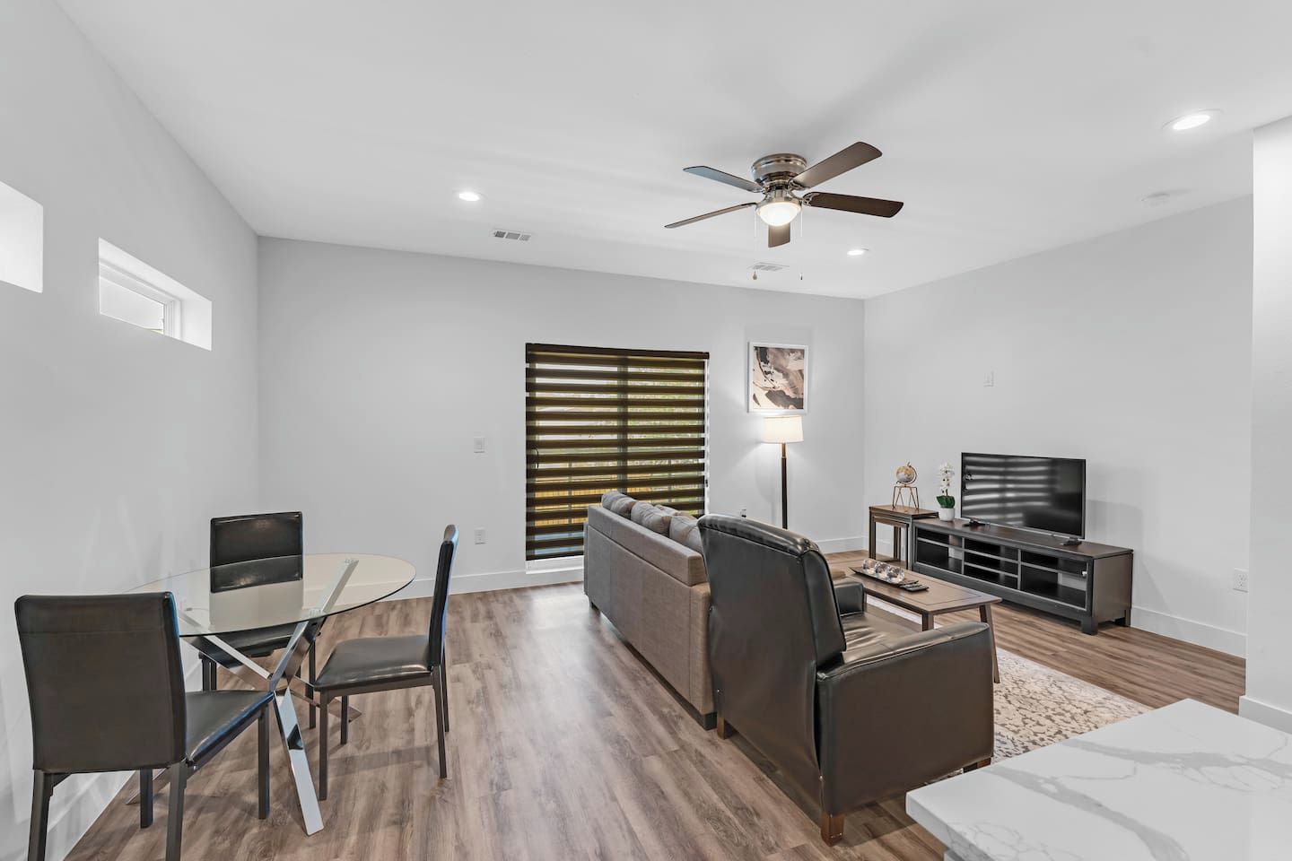 Step into a spacious, light-filled open concept living room seamlessly connected to a modern kitchen and cozy dining nook. Featuring plush seating, a large smart TV for streaming, a sleek glass-top table for four, and chic wood-grain flooring through