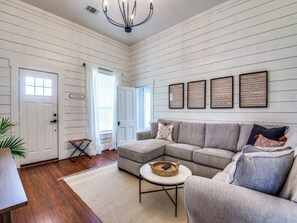 Sunlit shiplap living room with original hardwood floors, gray sectional, round coffee table, and large windows. Smart TV and high-speed Wi-Fi included. Modern farmhouse Airbnb in downtown Aubrey TX near Denton County wedding venues.